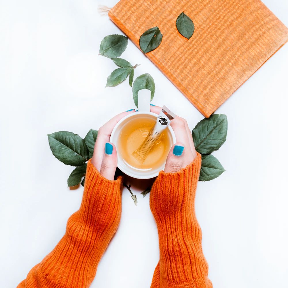Manos con suéter naranja sosteniendo una taza con un Tea Stick de Bistrotea